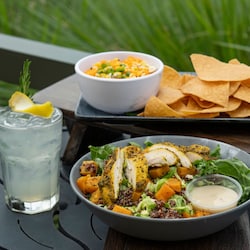 A bowl with chicken and sweet potatoes on top of lettuce next to a drink and a platter of tortilla chips and corn dip